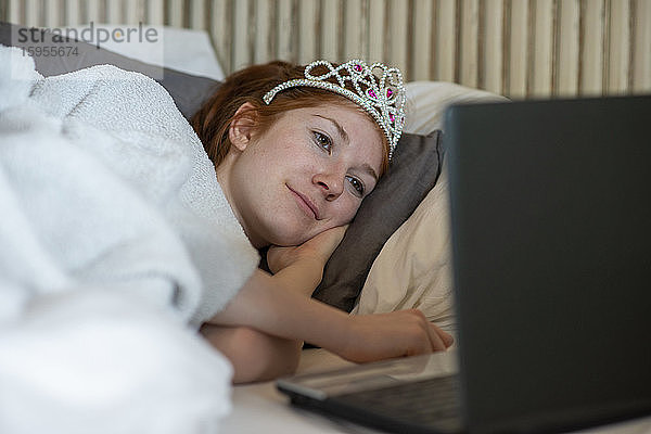 Porträt einer Teenagerin mit Diadem  die im Bett liegt und mit einem Laptop fernsieht