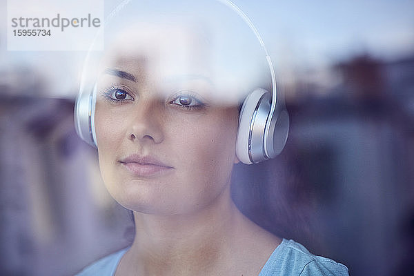 Porträt einer jungen Frau  die mit Kopfhörern Musik hört und aus dem Fenster schaut