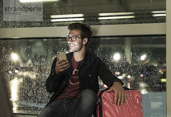Lächelnder junger Mann mit Koffer sitzt am verregneten Fenster und benutzt ein Smartphone