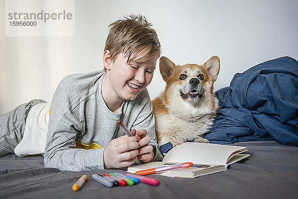Porträt eines glücklichen Jungen  der mit Filzstiften und Skizzenbuch mit seinem Hund auf dem Bett liegt