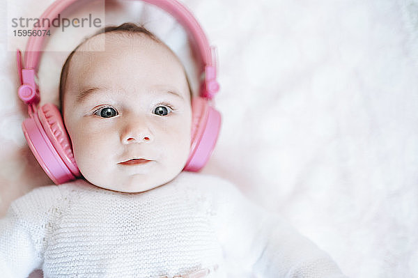 Porträt eines kleinen Mädchens mit übergrossen rosa Kopfhörern auf einer weissen Decke liegend