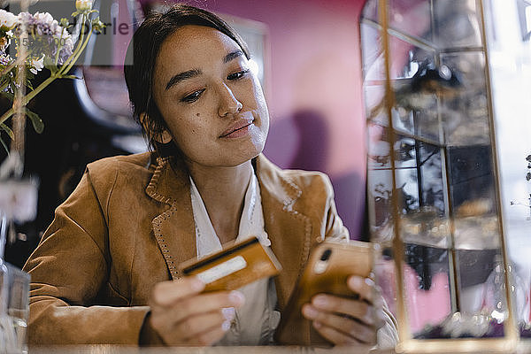 Nachdenkliche Frau betrachtet Smartphone und Kreditkarte im Second-Hand-Laden