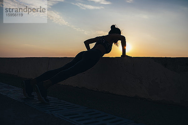 Ganzflächige Silhouette einer Athletin  die bei Sonnenaufgang an der Promenade Liegestützen macht