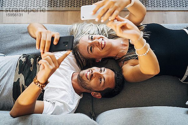 Porträt eines lachenden Paares  das auf der Couch liegt und sich mit Smartphones fotografiert