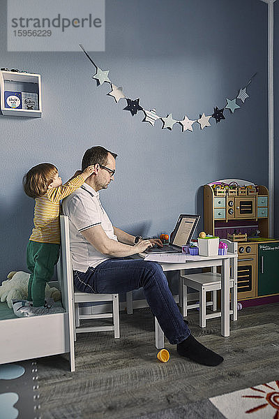 Mann arbeitet und benutzt Laptop im Kinderzimmer mit gleichzeitiger Kinderbetreuung