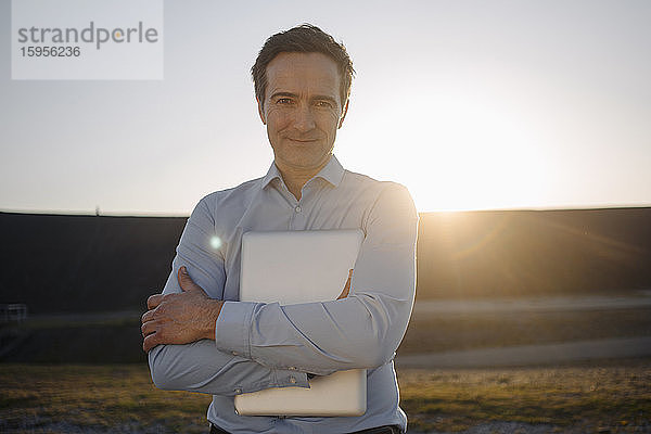 Porträt eines reifen Geschäftsmannes  der bei Sonnenuntergang einen Laptop auf einer stillgelegten Minenhalde hält