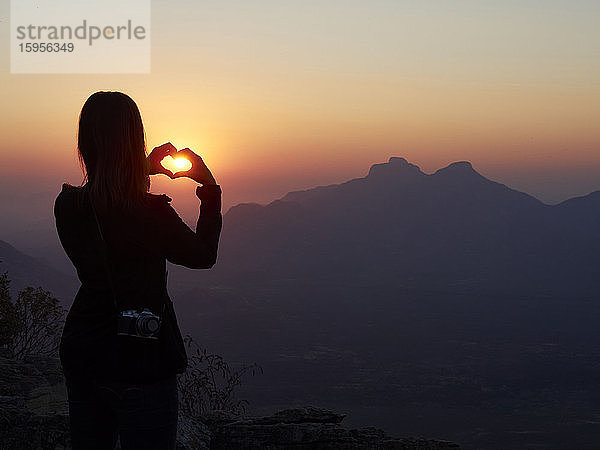 Silhouette einer Frau  die bei Sonnenuntergang mit ihren Händen das Herz formt  Serra da Leba  Angola