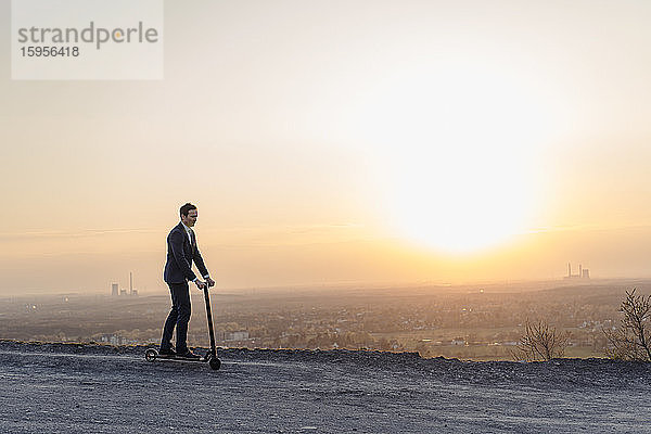 Ein reifer Geschäftsmann fährt bei Sonnenuntergang auf einem Kickroller auf einer stillgelegten Minenhalde
