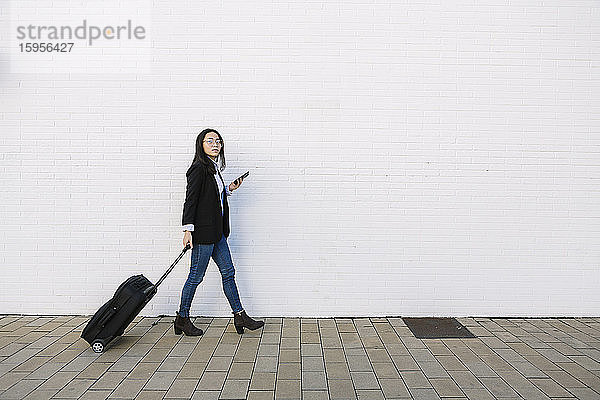 Junge Geschäftsfrau mit Mobiltelefon und Rollkoffer vor weißer Wand