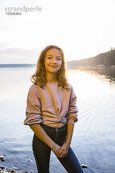 Lächelndes Mädchen steht bei Sonnenuntergang am Seeufer gegen den Himmel