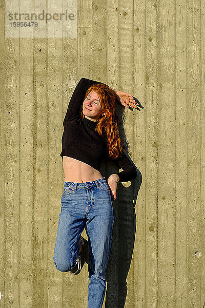 Porträt eines lächelnden Teenager-Mädchens mit geschlossenen Augen  das an einer Betonwand lehnt und das Sonnenlicht genießt