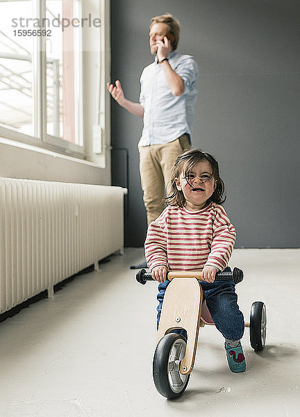 Weinendes Mädchen auf Balance-Fahrrad mit Vater am Telefon im Hintergrund