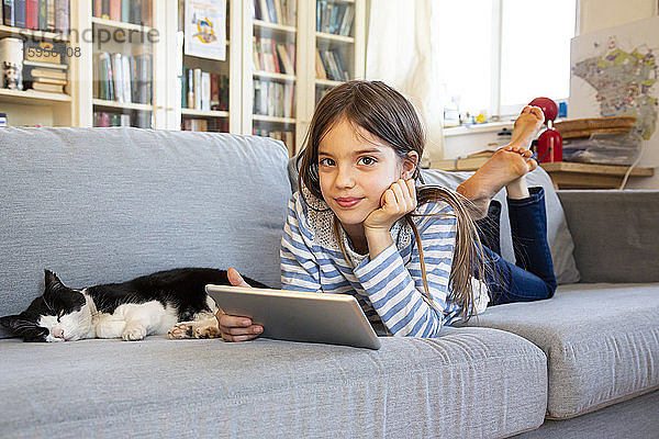 Porträt eines lächelnden Mädchens auf einer Couch liegend mit Katze und digitalem Tablett
