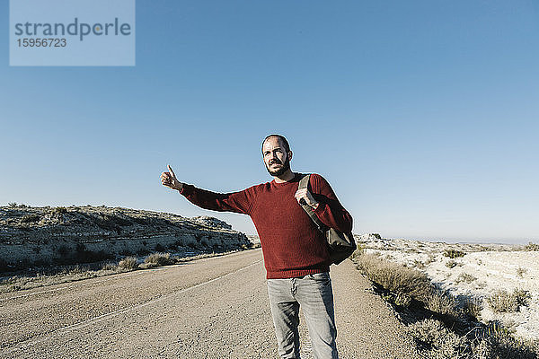 Mann gestikuliert beim Trampen am Straßenrand in der Wüste bei strahlend blauem Himmel