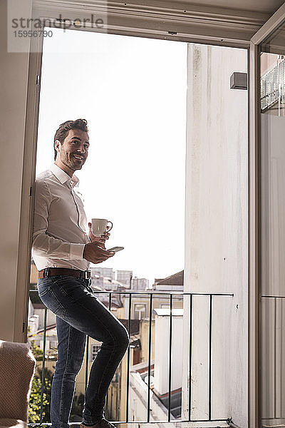 Junger Mann mit Handy und Kaffeetasse auf einem Balkon