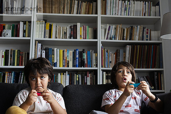 Jungen spielen Videospiel  während sie auf dem Sofa gegen ein Bücherregal im heimischen Wohnzimmer sitzen