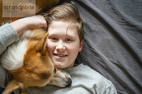 Porträt eines lächelnden Jungen  der auf dem Bett liegt und seinen Hund kuschelt