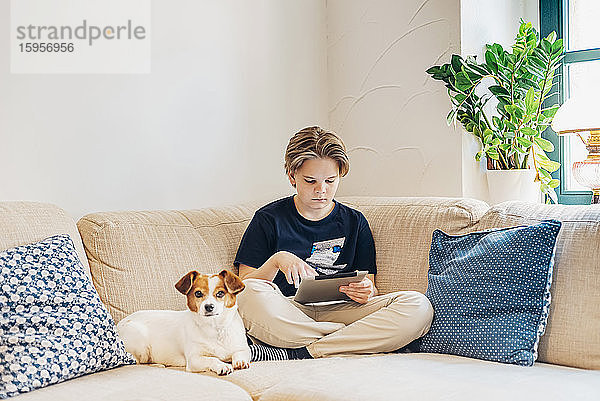 Junge mit Hund und Tablett auf der Couch sitzend im Wohnzimmer zu Hause