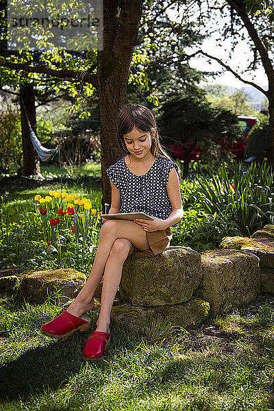 Lächelndes Mädchen sitzt im Garten und schaut auf das digitale Tablet