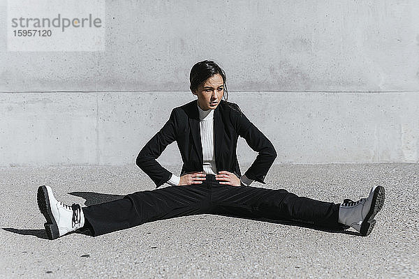 Junge Frau im schwarzen Anzug sitzt auf dem Boden vor einer Betonwand