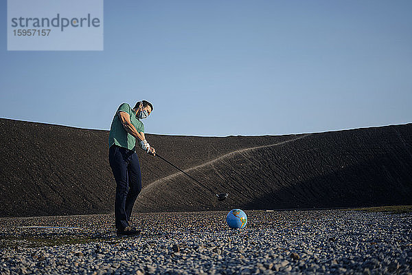 Reifer Mann mit Maske spielt Golf mit einem Globus auf einer stillgelegten Minenspitze