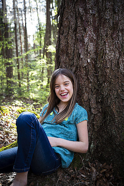 Porträt eines lachenden Mädchens  das sich im Wald an einen Baumstamm lehnt
