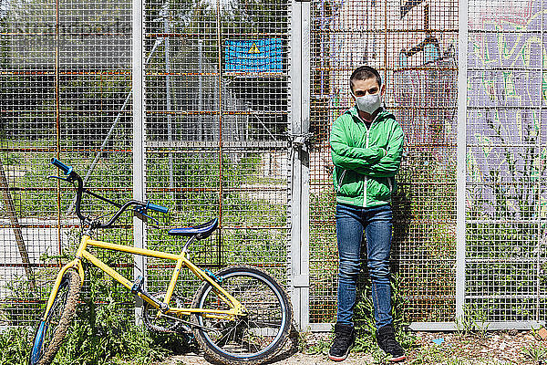 Porträt eines vorpubertären Jungen  der eine Gesichtsmaske trägt  während er mit verschränkten Armen mit dem Fahrrad gegen den Zaun steht