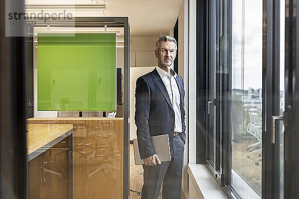 Porträt eines selbstbewussten reifen Geschäftsmannes mit Tablett im Büro am Fenster stehend