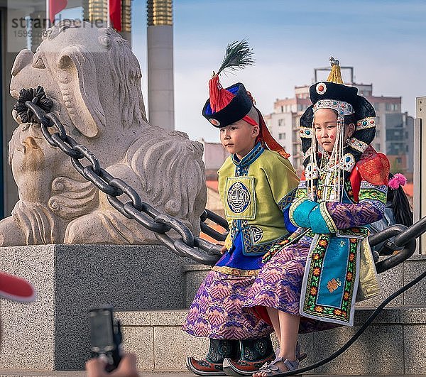 Mongolischer Junge in der traditionellen  traditionellen  traditionellen  traditionellen  traditionellen  traditionellen Tracht eines Mannes und eines Mädchens in der traditionellen Tracht einer Frau  die an einer Kette neben der Löwenstatue auf dem zentralen Platz sitzen  Hauptstadt von Ulaanbaatar  Mongolei  Asien