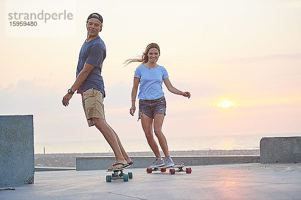 Ein junges Paar  Mann und Frau  skateboarden bei Sonnenuntergang am Strand.