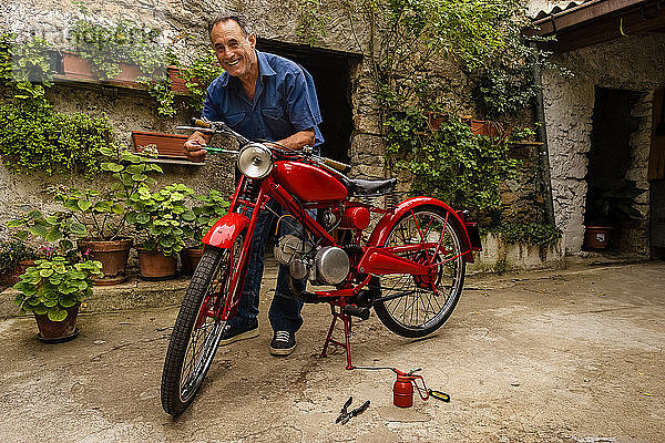 Ein reifer Mann steht im Hof und repariert ein rotes Oldtimer-Motorrad.