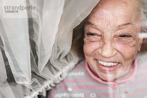 Lächelnde alte Frau  die während der Coronavirus-Quarantäne durch ein Fenster schaut.