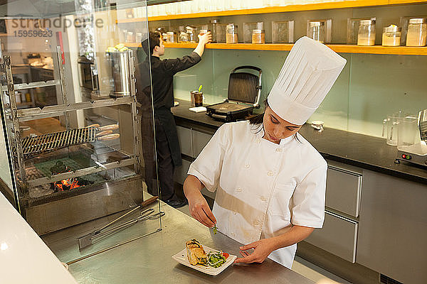 Köchin mit Kochmütze steht an der Arbeitsplatte in der Großküche und garniert den Teller mit Lebensmitteln.