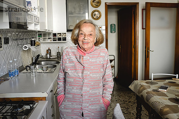 Alte Frau steht während der Coronavirus-Quarantäne in ihrer Küche und lächelt in die Kamera.