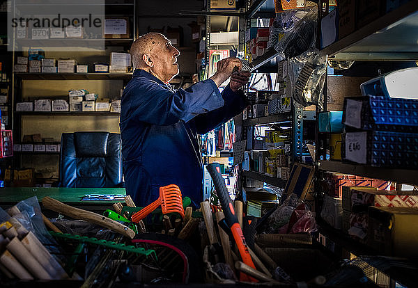 Ein Eisenwarenhändler  ein älterer Mann in einem Overall  der Regale mit nützlichen Haushaltsgegenständen und Werkzeugen durchsucht.