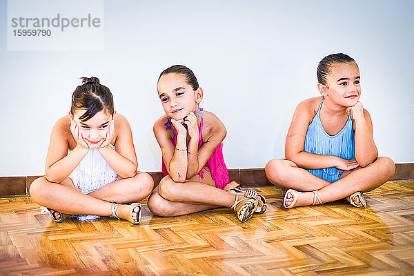 Drei junge Mädchen im Tanzkurs  geschminkt und in Sommerkleidern auf Parkett sitzend.