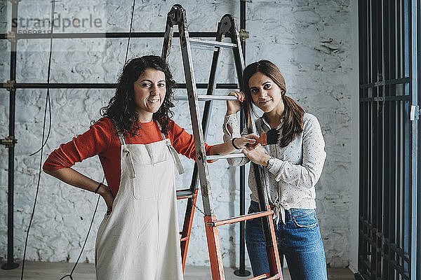Zwei mittelgroße erwachsene Frauen  die sich in ihrem neuen Laden auf Trittleitern mit Pinsel abstützen  Porträt
