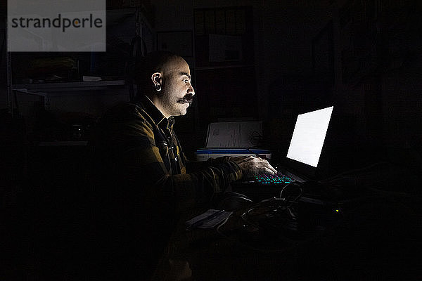 Kahlköpfiger Mann mit Schnurrbart sitzt in einem dunklen Raum  tippt am Laptop und isoliert sich während der Corona-Krise selbst.