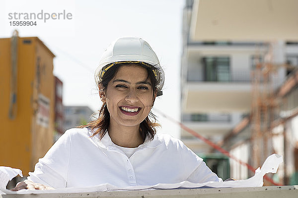 Weibliche Architektin mit weißem Schutzhelm  die auf der Baustelle arbeitet und in die Kamera lächelt.