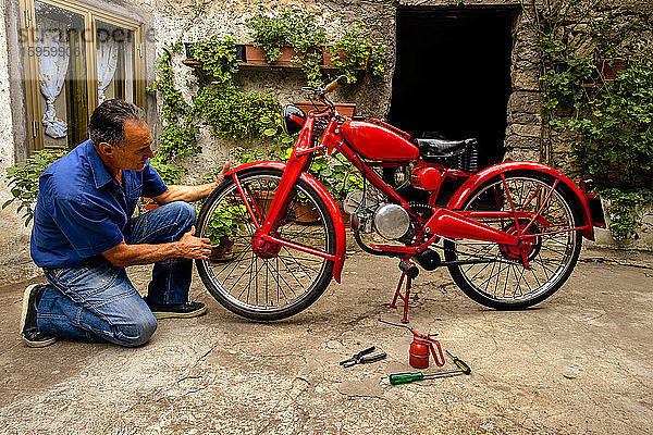 Ein reifer Mann kniet im Hof und repariert ein rotes Oldtimer-Motorrad.