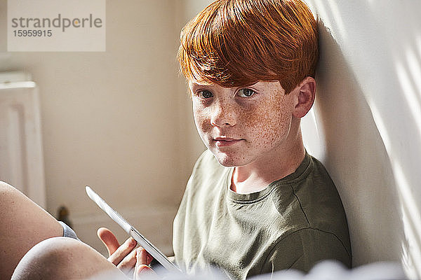 Junge mit roten Haaren sitzt auf dem Boden in einem sonnigen Raum und hält ein digitales Tablett in der Hand.