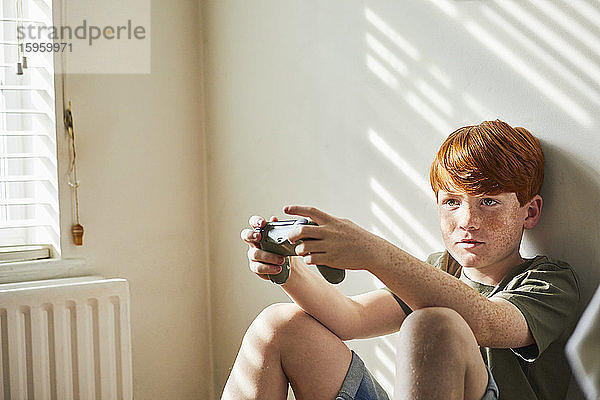 Junge mit roten Haaren  der in einem sonnigen Raum auf dem Boden sitzt und einen Spielkonsolen-Controller in der Hand hält.