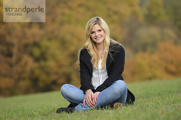 Lächelnde blonde junge Frau sitzt auf herbstlicher Wiese