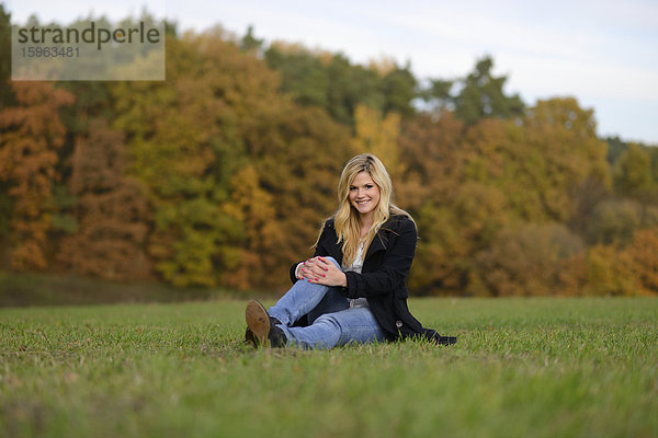 Lächelnde blonde junge Frau sitzt auf herbstlicher Wiese