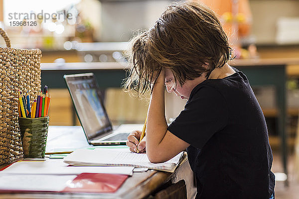 6-jähriger Junge  der eine Fernschulung mit seinem Lehrer hat und sich dabei auf das Schreiben konzentriert.