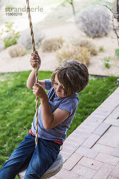 Sechsjähriger Junge  der mit einer Seilschaukel auf einer breiten Veranda im Schatten schaukelt.