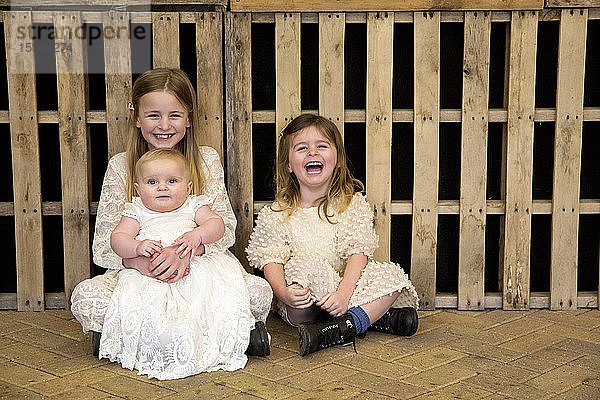 Porträt von drei jungen Schwestern während der Taufzeremonie in einer historischen Scheune  die auf dem Boden sitzen und in die Kamera lächeln.