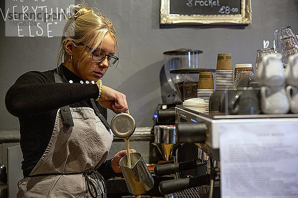 Blonde Frau mit Brille und Schürze  die an einer Espressomaschine in einem Café steht und Milch in eine Metallkanne gießt.