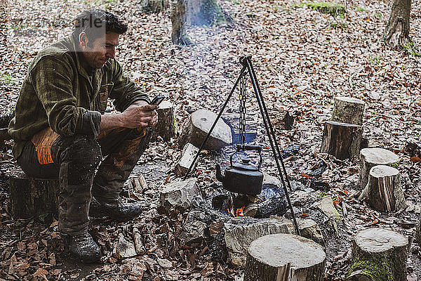 Mann sitzt an einem Lagerfeuer im Wald und kocht einen Wasserkessel.