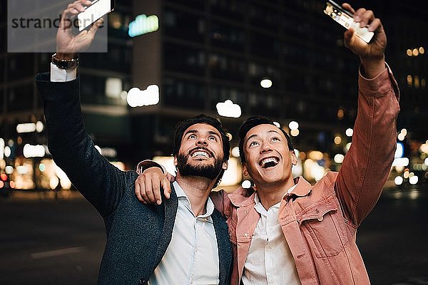 Glücklicher Mann und männlicher Freund schauen mit Smartphones auf  während sie nachts in der Stadt gestikulieren